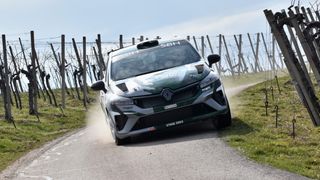 Ein Rallye-Auto fährt auf einer unbefestigten Straße mit einem grün-weißen Farbschema, 'STAGE ZERO' steht auf der vorderen Stoßstange. Dahinter befinden sich Holzzäune und ein Zaun.