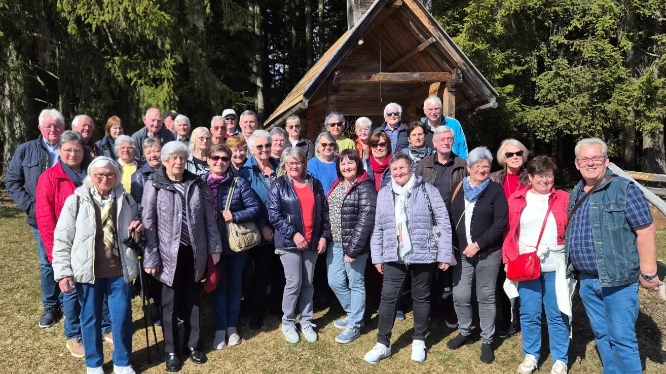 Eine Gruppe älterer Personen posiert für ein Foto vor einer Holzstruktur, umgeben von Bäumen. Einige tragen Brillen, Schals und Jacken.