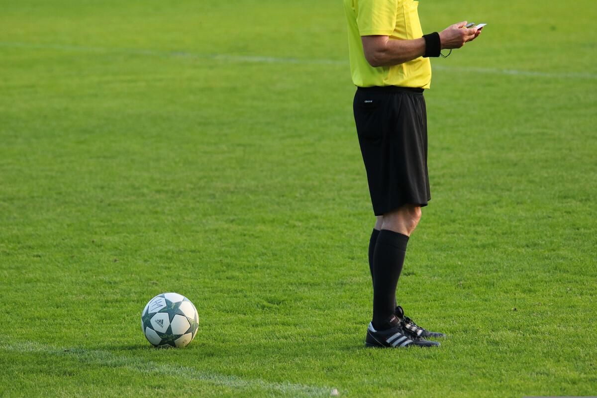A soccer referee stands on a field, holding a whistle and a card. A soccer ball is nearby, and the field has white lines.