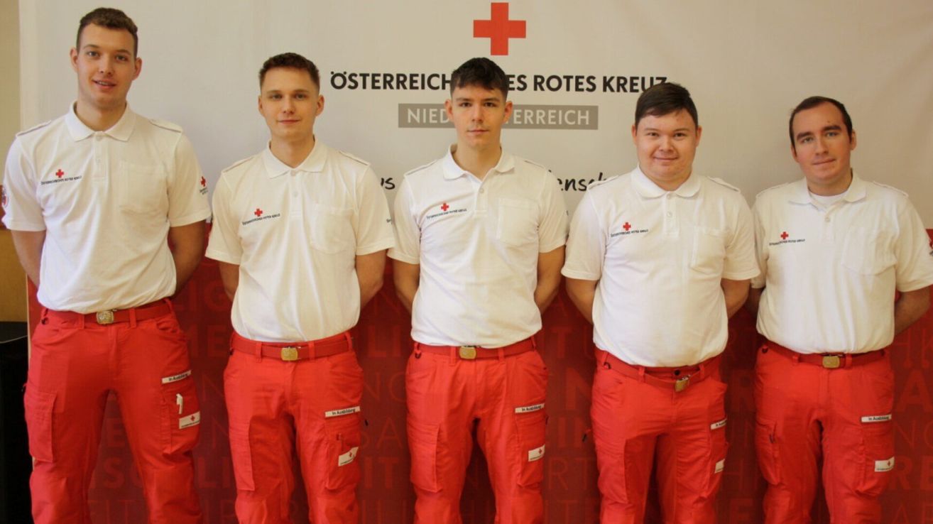Three individuals in white polo shirts and red trousers stand next to each other in front of a white wall with red crosses and text.