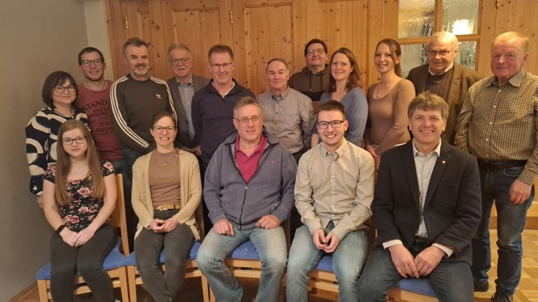 Eine Gruppe von Menschen versammelt sich in einem Raum mit Holzwänden und lächelt für ein Foto. Sie tragen Freizeitkleidung, einige mit Brillen. Im Hintergrund befinden sich Stühle und eine Holztür.