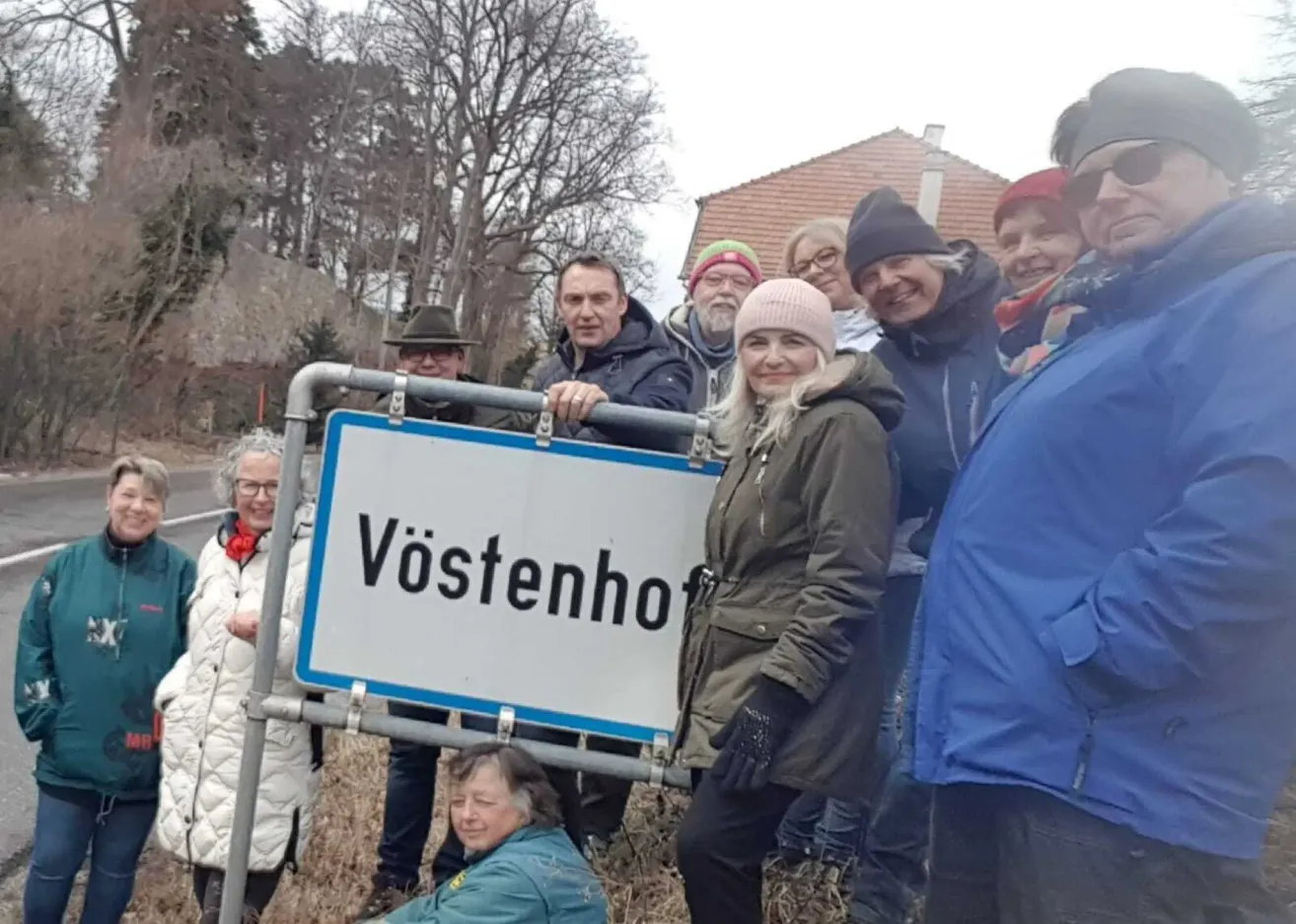 Eine Gruppe von Menschen steht um ein weißes Schild mit dem Namen Vöstenho. Sie tragen alle Winterkleidung. Hinter ihnen steht ein Haus mit einer Ziegelwand. Im Hintergrund befinden sich Bäume und eine Straße.