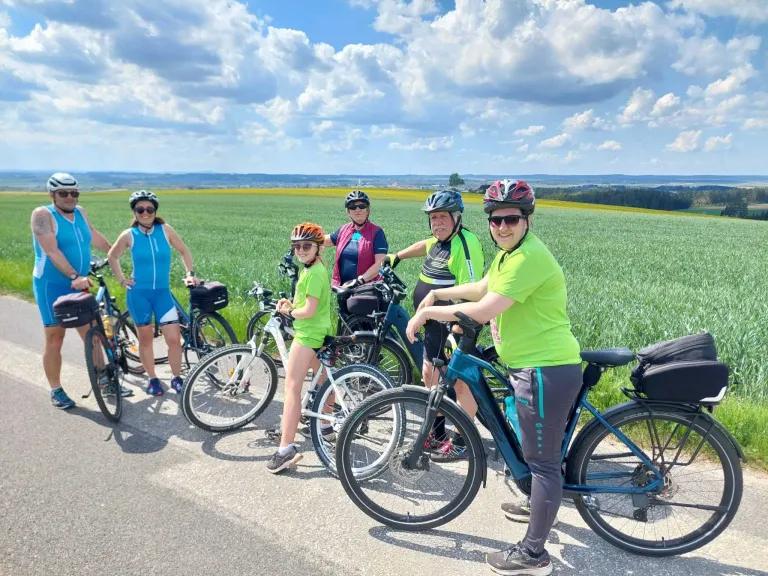 A group of cyclists in green shirts and helmets pose with their bikes on a sunny day, standing in a field with a clear sky and distant mountains.