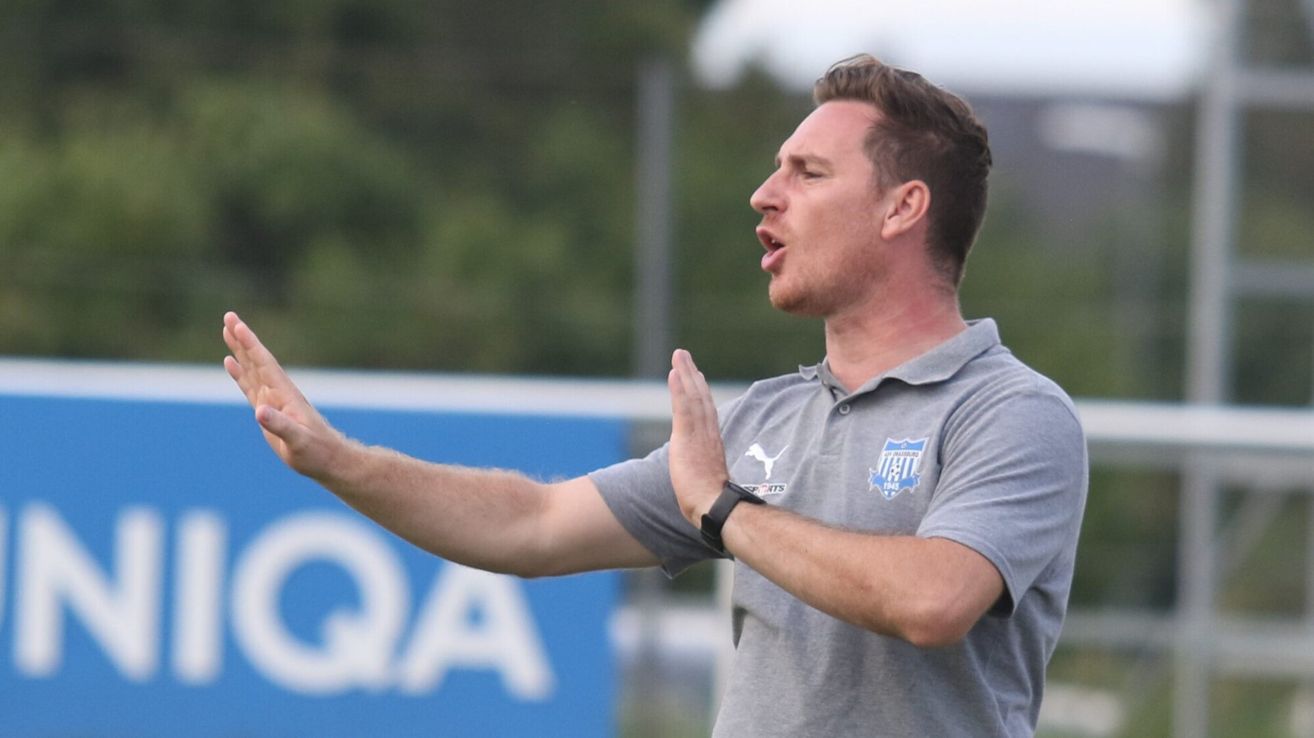 Ein Mann in einem grauen Shirt macht Gesten mit seinen Händen auf einem Feld, möglicherweise als Trainer. Hinter ihm befindet sich ein blauer Banner mit weißen Buchstaben.