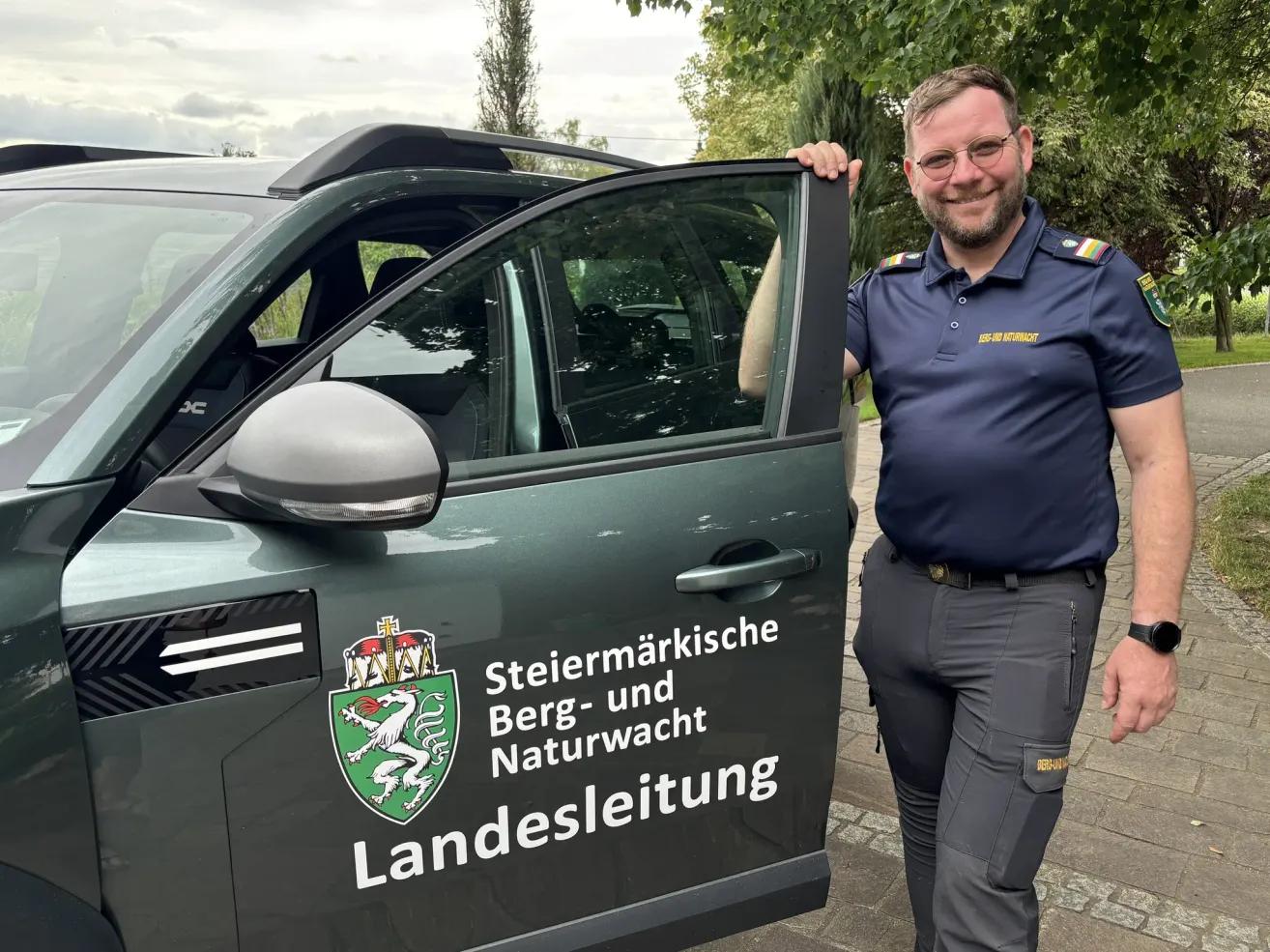 A man stands next to a dark green vehicle, smiling. The vehicle has a logo and the words 'Steiermarkische Berg- und Naturschutz' on its side. The man is wearing a blue shirt and gray pants.