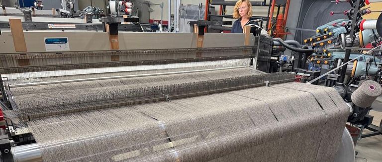 Eine Frau mit Brille steht vor einer großen Textilmaschine und beobachtet die Produktion von grauem Stoff.