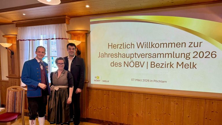 Ein Mann und eine Frau stehen lächelnd vor einer Projektionsleinwand, während sie ein Foto machen. Die Leinwand zeigt die Worte 'Herzlich Willkommen Jahreshauptversammlung des NOBV | Bezirk 07. März 2026 in Pöchlarn'.