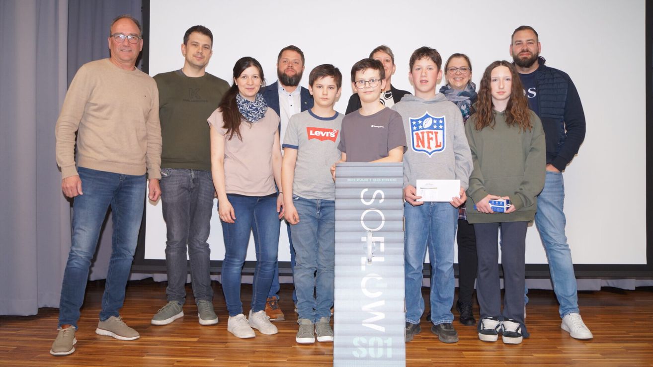 Eine Gruppe von Menschen steht auf einem Holzboden. Einige sind in Freizeitkleidung gekleidet, während andere formell gekleidet sind. Sie alle lächeln für ein Foto. Ein Banner mit dem Text 'SOS' steht in der Mitte. Einige halten Urkunden in den Händen.