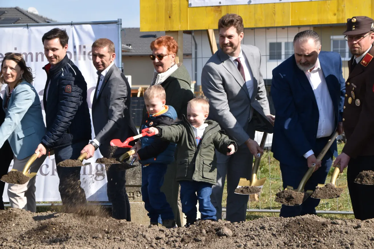 Eine Gruppe von Menschen, einschließlich Erwachsener und Kindern, stehen um einen Haufen Erde und halten Schaufeln, möglicherweise für eine Grundsteinlegungszeremonie. Hinter ihnen befindet sich ein Gebäude mit einem gelben Schild und einem Zaun.