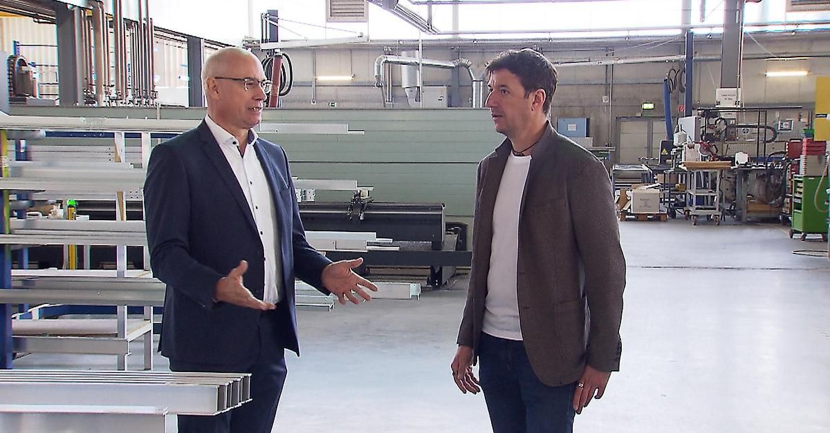 Zwei Männer in Geschäftskleidung stehen in einer Fabrik, einer gestikuliert, während der andere zuhört. Maschinen und Geräte umgeben sie.