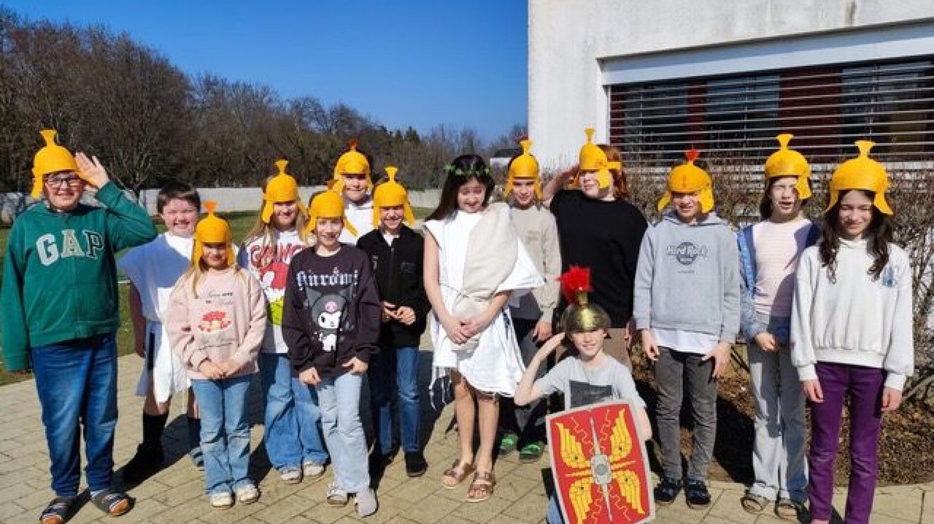 Eine Gruppe von Kindern, die als Römer gekleidet sind, steht vor einem Gebäude. Einige tragen Helme und einer hält einen Schild. Sie sind in Freizeitkleidung und Bäume sind im Hintergrund.