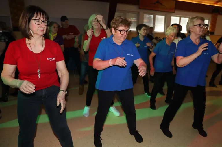 Eine Gruppe von Frauen, einige in roten und einige in blauen Shirts, tanzt in einem Raum mit grüner Beleuchtung auf dem Boden.