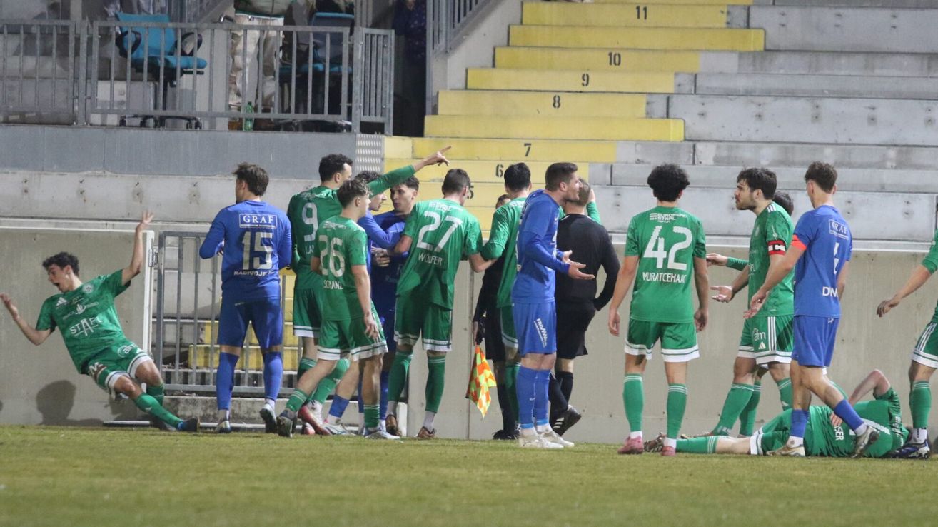 Fußballspieler in grünen Trikots stehen gedrängt zusammen, ein Schiedsrichter hält eine Flagge. Sie sind auf einem Feld mit nummerierten Tribünen im Hintergrund.