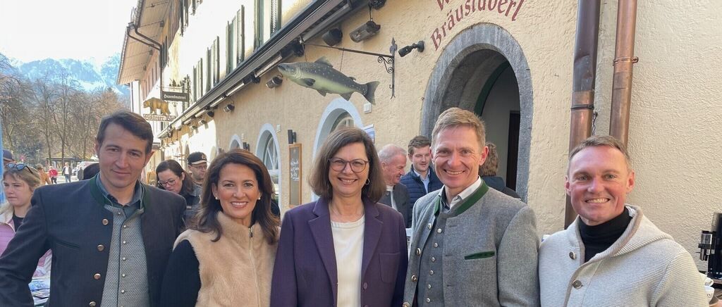 Five people stand outside a building with the name Braustüberl. They are smiling for a photo. Behind them are more people. Above the entrance is a fish sign.