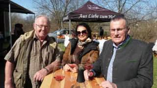 Three individuals, two men and a woman, are seated at a wooden table, posing for a photograph. They each have a glass of wine, and the woman is wearing sunglasses. Behind them, a tent with the text 'Aus Liebe zum Hof' is visible, along with a car and a person.