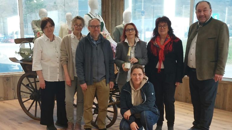 Eine Gruppe von Menschen steht vor einer Glaswand mit Mannequins und posiert für ein Foto. Eine Frau hält eine Flasche, während andere Brillen und Jacken tragen.