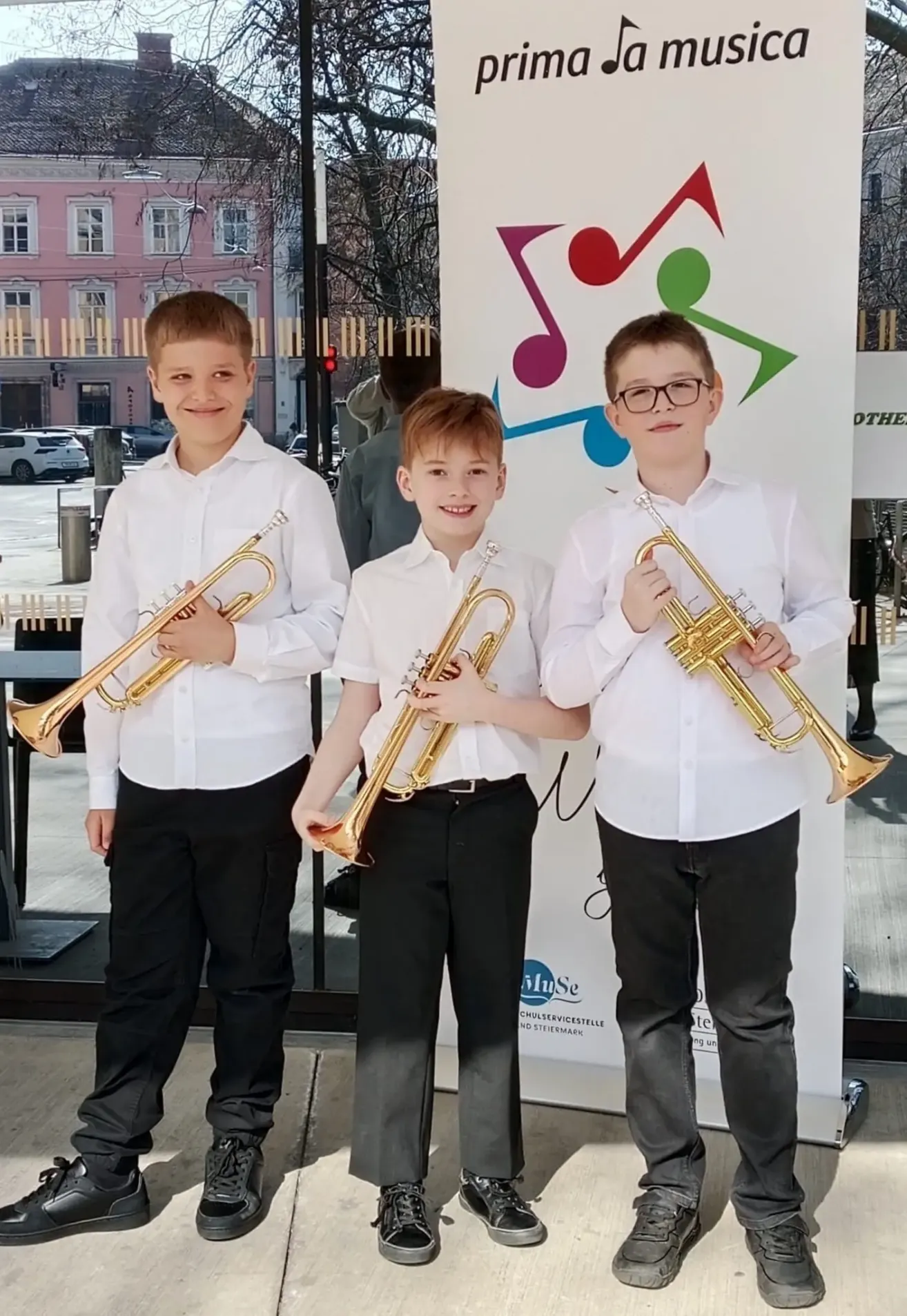 Three boys in matching outfits stand before a colorful banner, each holding a trumpet. They are smiling and appear to be posing for a photograph.