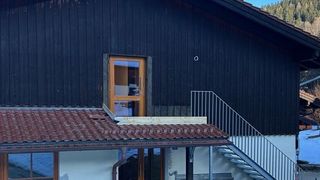 A modern house with a black exterior, brown roof, glass door, and metal stairs. The roof has a drain pipe, and the walls are wooden. Snow is visible in the background.
