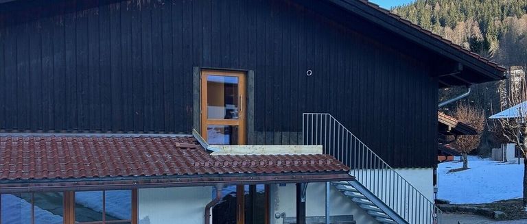 A modern house with a black exterior, brown roof, glass door, and metal stairs. The roof has a drain pipe, and the walls are wooden. Snow is visible in the background.
