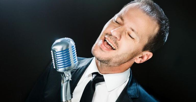 Ein Mann in Anzug und Krawatte singt in ein Mikrofon, die Augen geschlossen und der Mund offen.