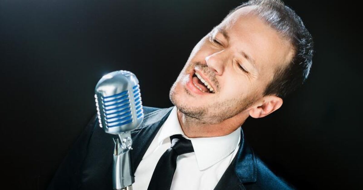 Ein Mann in Anzug und Krawatte singt in ein Mikrofon, die Augen geschlossen und der Mund offen.