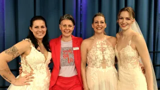 Four women smile for a photo. One wears a red blazer over a printed t-shirt, while the others wear white dresses. They stand in front of a blue curtain.