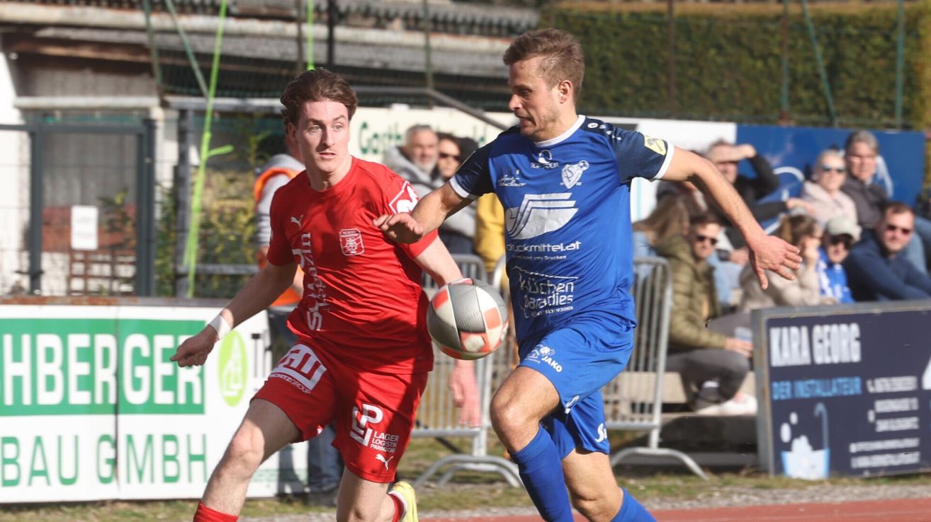 Zwei Fußballspieler in roten und blauen Trikots kämpfen auf einem Rasenfeld um den Ball, während eine Menge im Hintergrund zusieht.