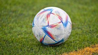 A soccer ball with a multicolored design is resting on a grassy field. The ball is white and blue with patches of red, yellow, and green.