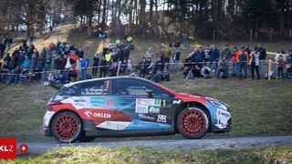 A rally car drives on a sloped road with spectators. The car is blue, white, and red with a Hyundai logo and sponsored by Orlen and other brands.