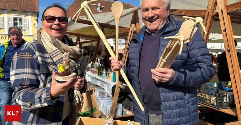 Ein Mann und eine Frau lächeln in die Kamera und halten Holzgegenstände. Die Frau hält ein kleines vogelartiges Objekt. Sie stehen auf einem Markt mit Ständen im Hintergrund.