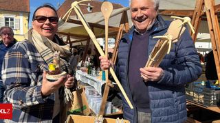Ein Mann und eine Frau lächeln in die Kamera und halten Holzgegenstände. Die Frau hält ein kleines vogelartiges Objekt. Sie stehen auf einem Markt mit Ständen im Hintergrund.