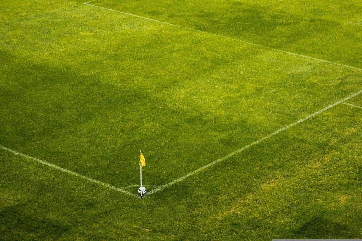 Eine Vogelperspektive eines Fußballfeldes mit weißen Begrenzungslinien und einer gelben Eckflagge. Das Feld ist grün und ein Fußball liegt an der Ecke.
