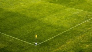 An aerial view of a soccer field with white boundary lines and a yellow corner flag. The field is green, and a soccer ball is placed at the corner.