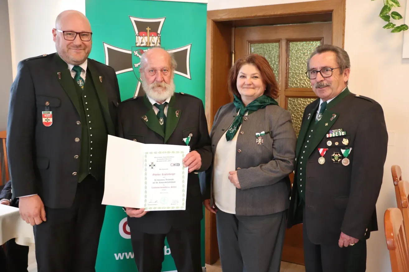 Four people dressed in formal attire stand before a green banner with a cross. A man holds a certificate, while a woman and two men stand beside him.