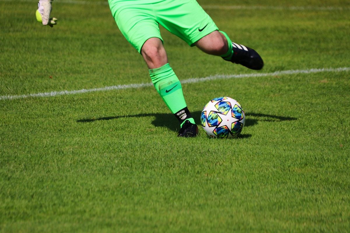 Ein Fußballspieler in grünen Uniformen und schwarzen Stollenschuhen tritt einen bunten Fußball auf einem Rasenfeld.