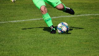 A soccer player in green uniforms and black cleats kicks a colorful soccer ball on a grassy field.