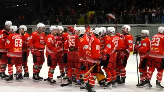A hockey team wearing red uniforms stands on the ice rink, celebrating a win. They are all wearing helmets and holding hockey sticks. Behind them, a crowd is watching.