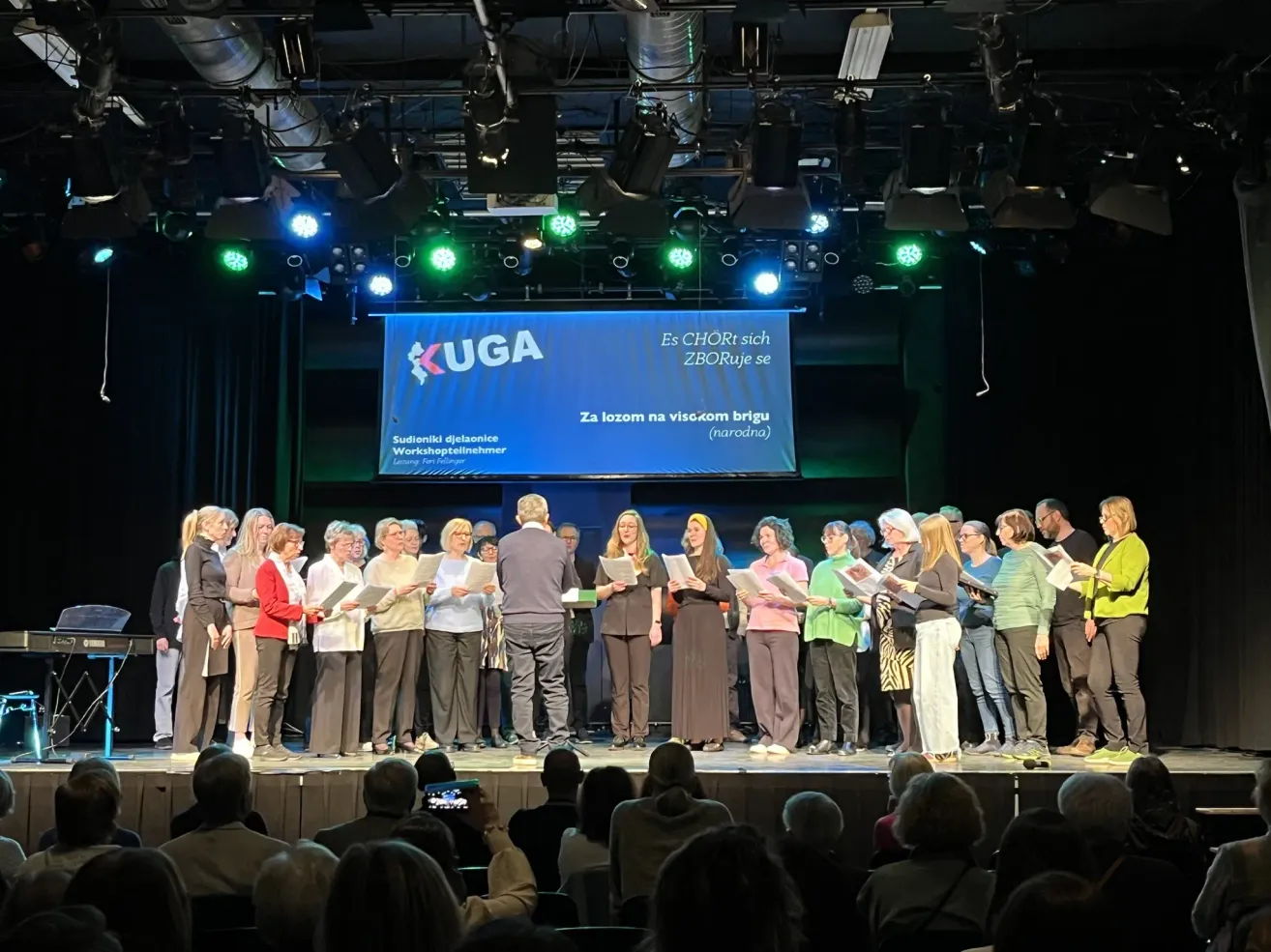 A choir performs on stage with a large banner reading 'KUGA' and 'Es CHORt sich ZBORuJe se'. Behind them, people hold papers and appear to be singing. Spotlights are on the ceiling.