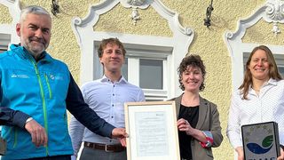 Four individuals stand in front of a building, two women and two men, smiling, with one man holding a framed certificate and another holding a framed sign.