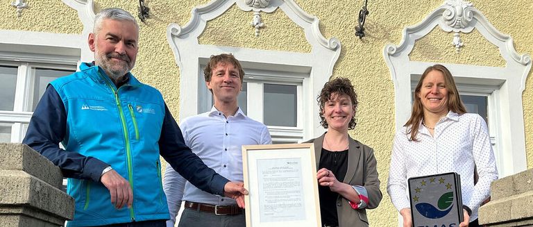 Four individuals stand in front of a building, two women and two men, smiling, with one man holding a framed certificate and another holding a framed sign.