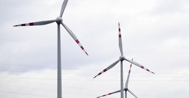Two wind turbines with red and white blades are positioned against a cloudy sky with electrical wires below.