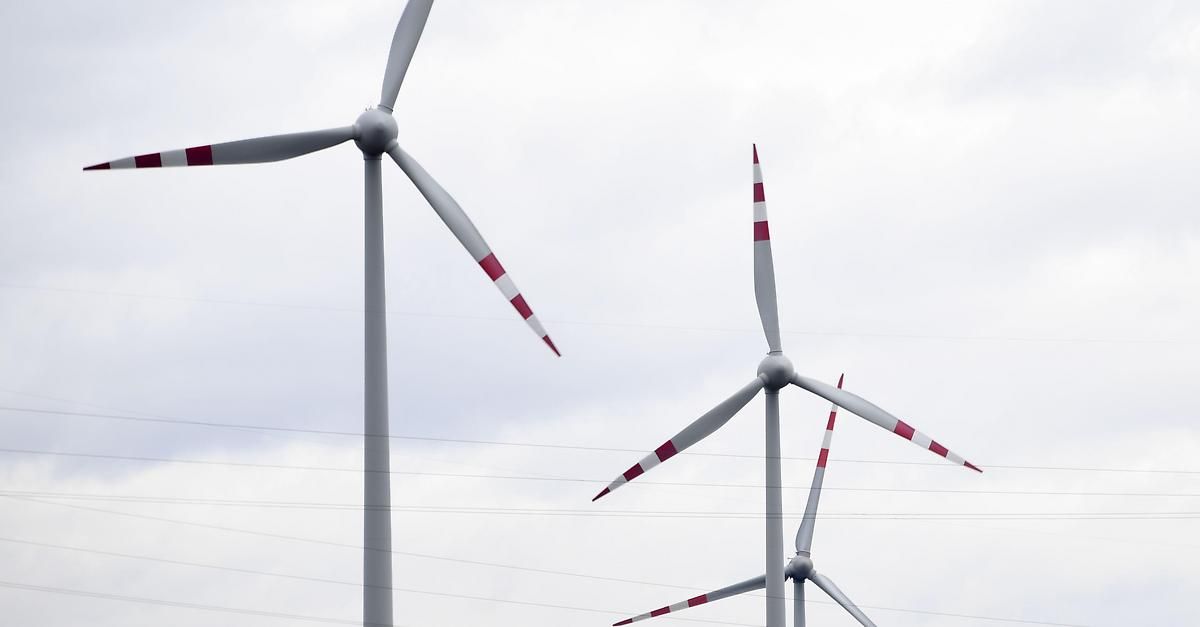 Zwei Windturbinen mit roten und weißen Rotorblättern stehen gegen einen bewölkten Himmel mit elektrischen Leitungen darunter.