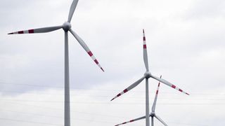 Two wind turbines with red and white blades are positioned against a cloudy sky with electrical wires below.