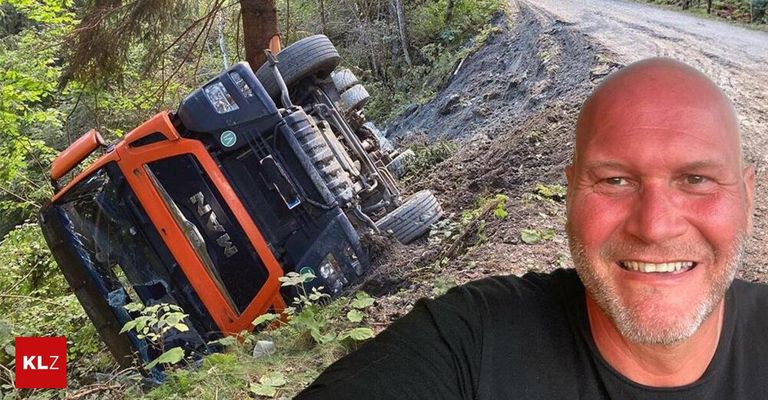 Ein großer orangefarbener und schwarzer Traktor ist auf einer schlammigen Straße umgestürzt, neben ihm steht ein lächelnder Mann.