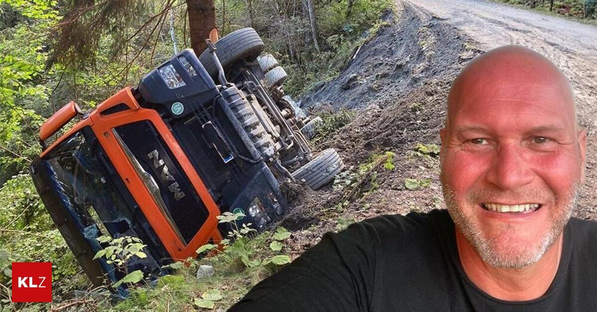 Ein großer orangefarbener und schwarzer Traktor ist auf einer schlammigen Straße umgestürzt, neben ihm steht ein lächelnder Mann.