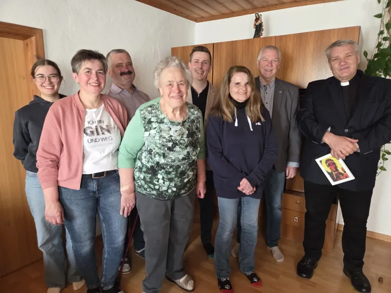 A group of people, including an elderly woman with a bandage on her arm, is standing in a room with wooden flooring. They are all smiling for the camera.