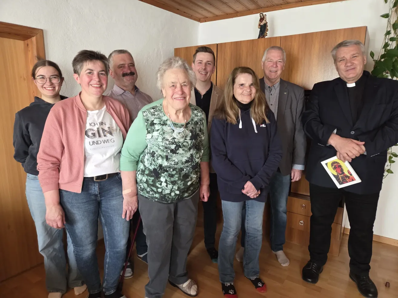 A group of people, including an elderly woman with a bandage on her arm, is standing in a room with wooden flooring. They are all smiling for the camera.