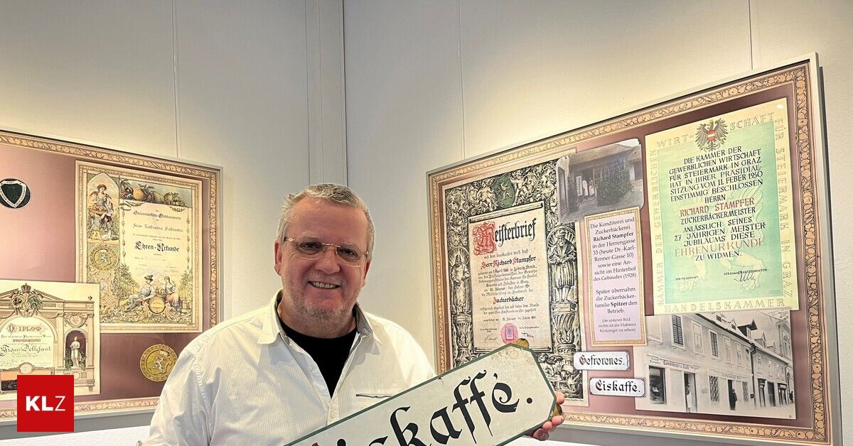 A man stands in front of a white wall holding a sign that reads 'kaffee'. Behind him are two framed documents with ornate designs.
