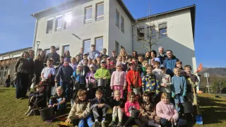 A group of children and adults pose for a photo in front of a building with a small tree, wearing winter clothes and holding buckets.