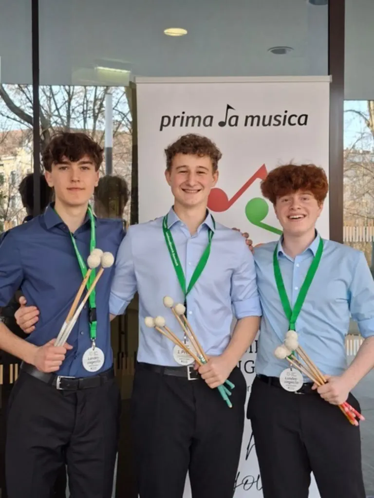 Three young men in blue shirts and black pants stand next to each other, each holding a set of drumsticks. They are standing in front of a white banner with the text 'prima Ja musica'.
